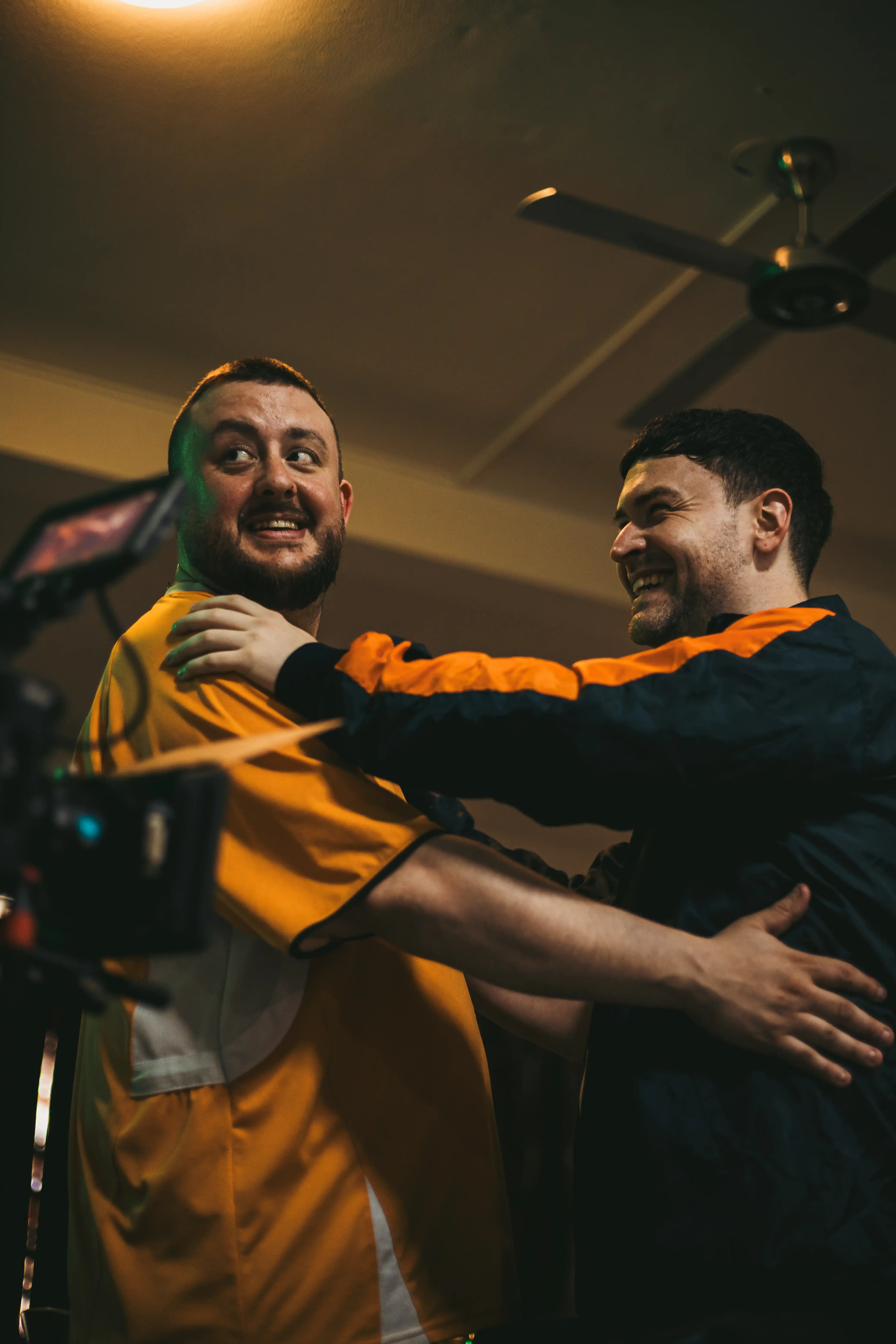 Behind the scenes picture of two men holding each other. A professional videography camera can be seen poking in the bottom left of the image.