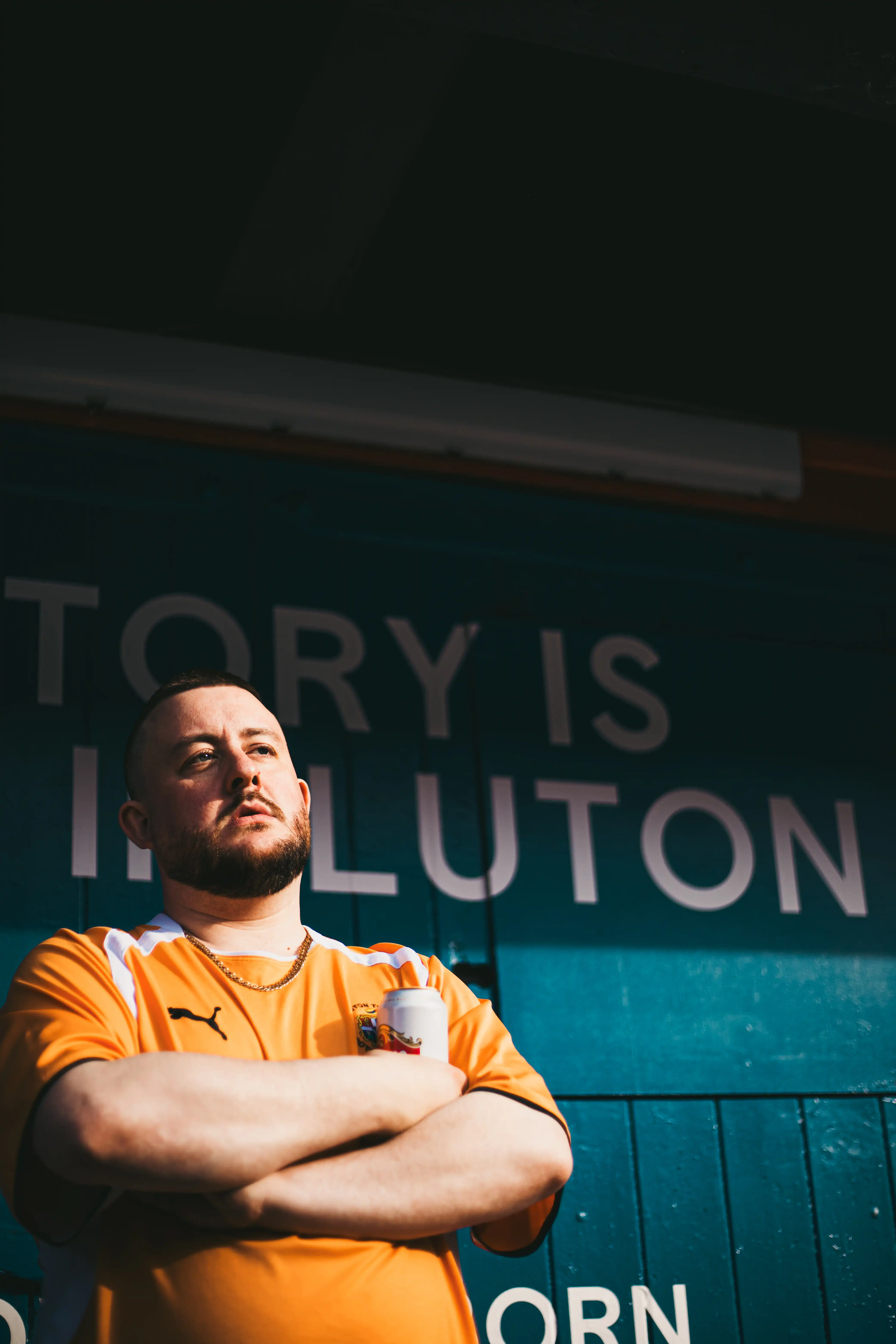 A man wearing a football shirt starring into the distance standing in front of a wooden wall painted blue.