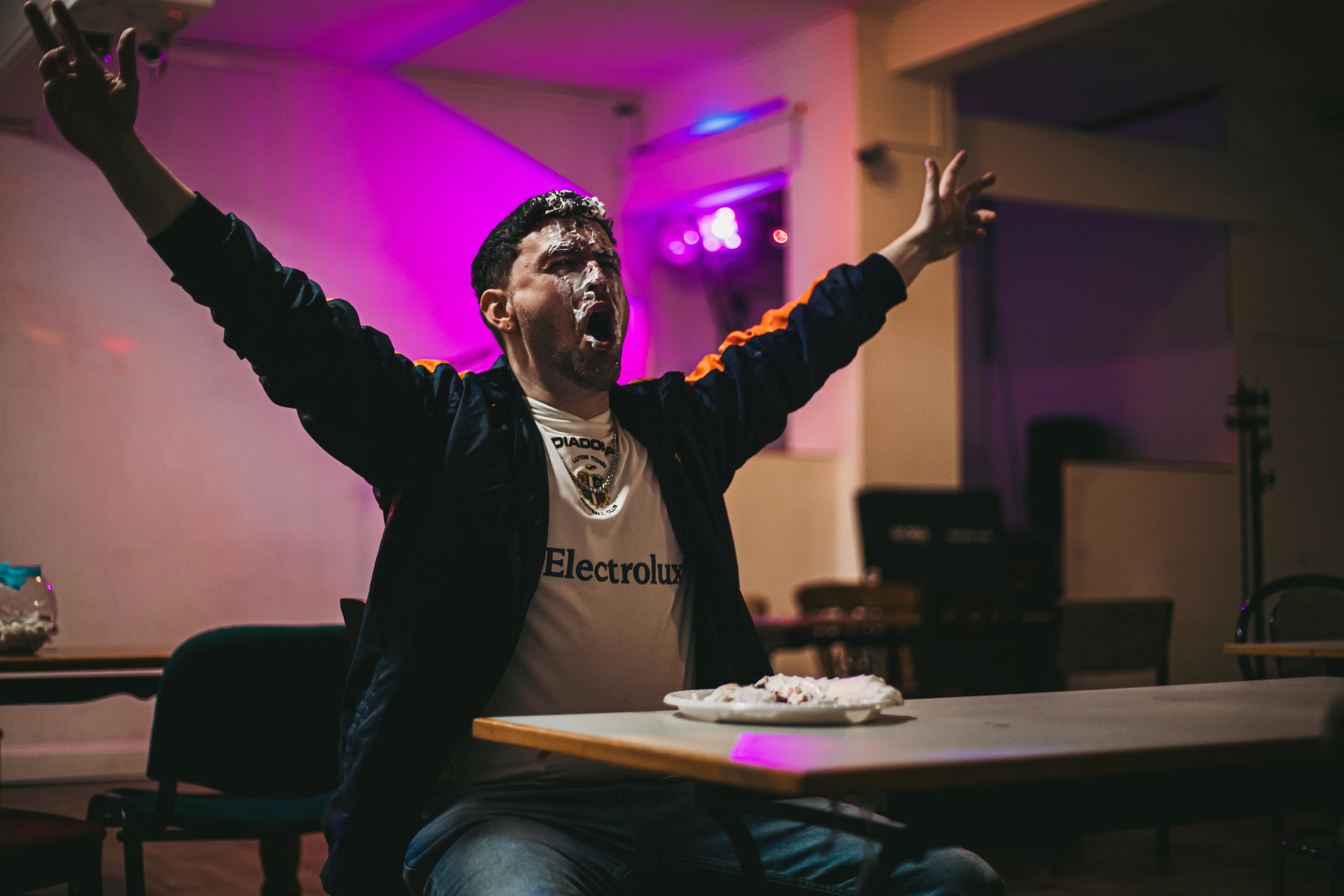 A man sitting alone at a table with whipped cream on his face. Sitting in front of a pie with his hands held in the air. Behind him is a bright purple light.