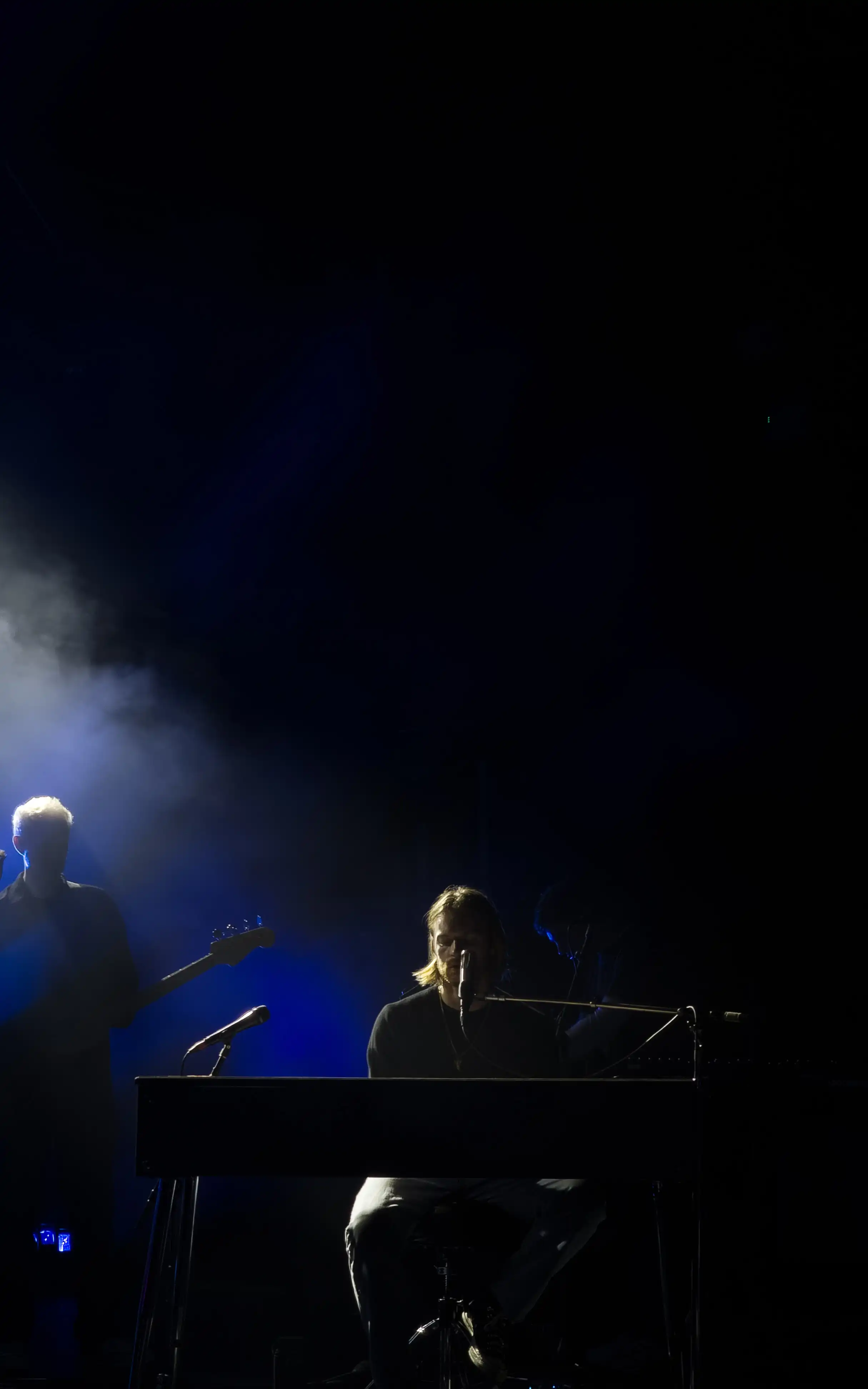 A dark and gloomy shot of Finneas playing piano on stage infront of a crowded audience.