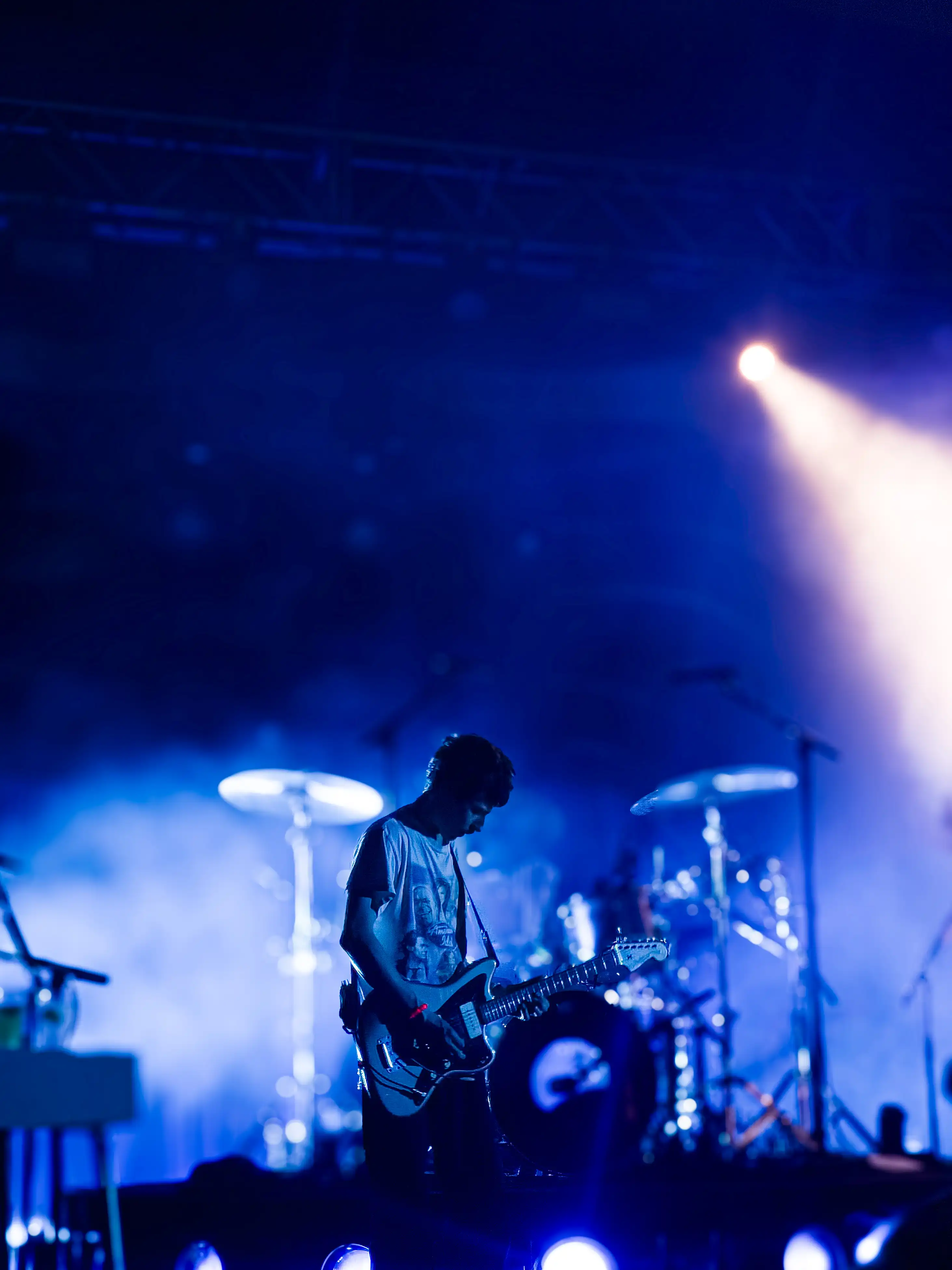 A dark and gloomy blue image of the guitarist for Finneas playing guitar on stage infront of a crowded audience.