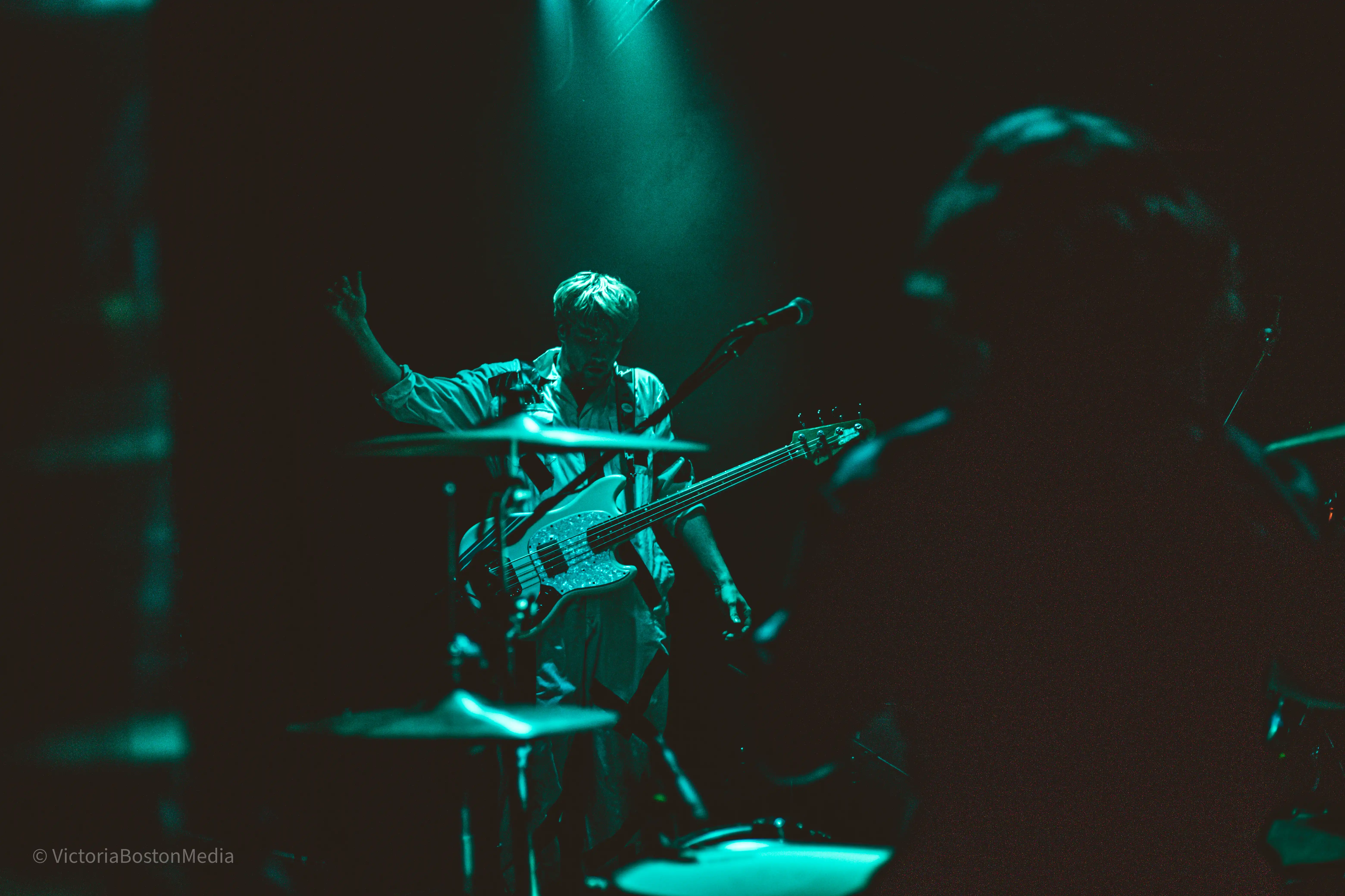 A bassist and a drummer facing each other on a dark and gloomy stage with a turquoise stage light above them.