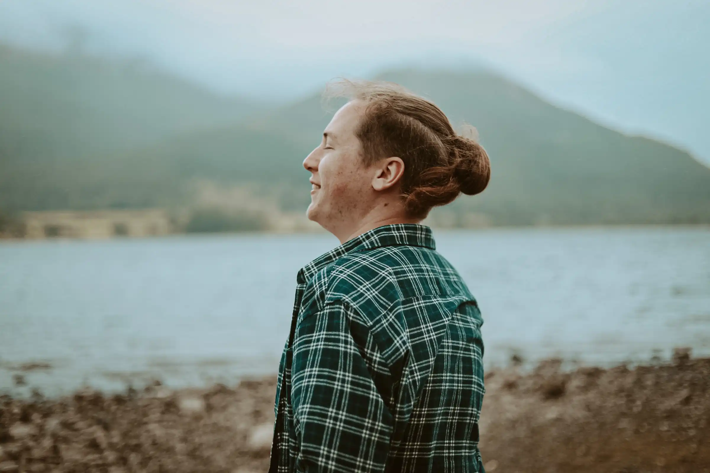 A portrait shot of a male with a top knot wearing a green plaid shirt looking off into the distance.