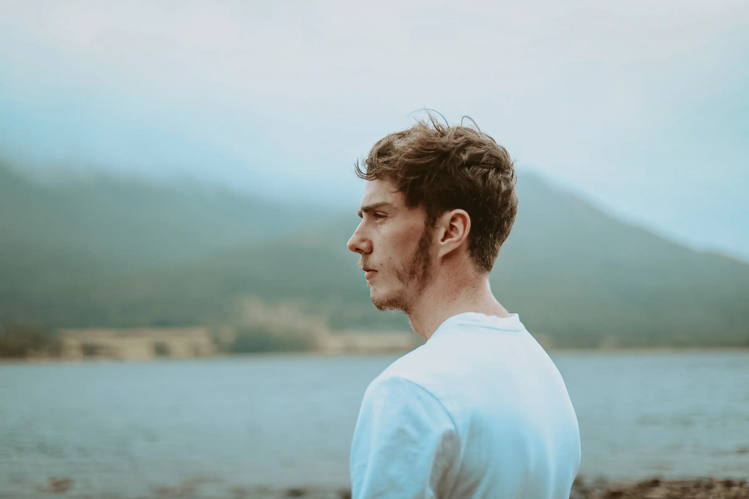 A portrait shot of a male standing in front of a lake wearing a white t-shirt, looking off into the distance.
