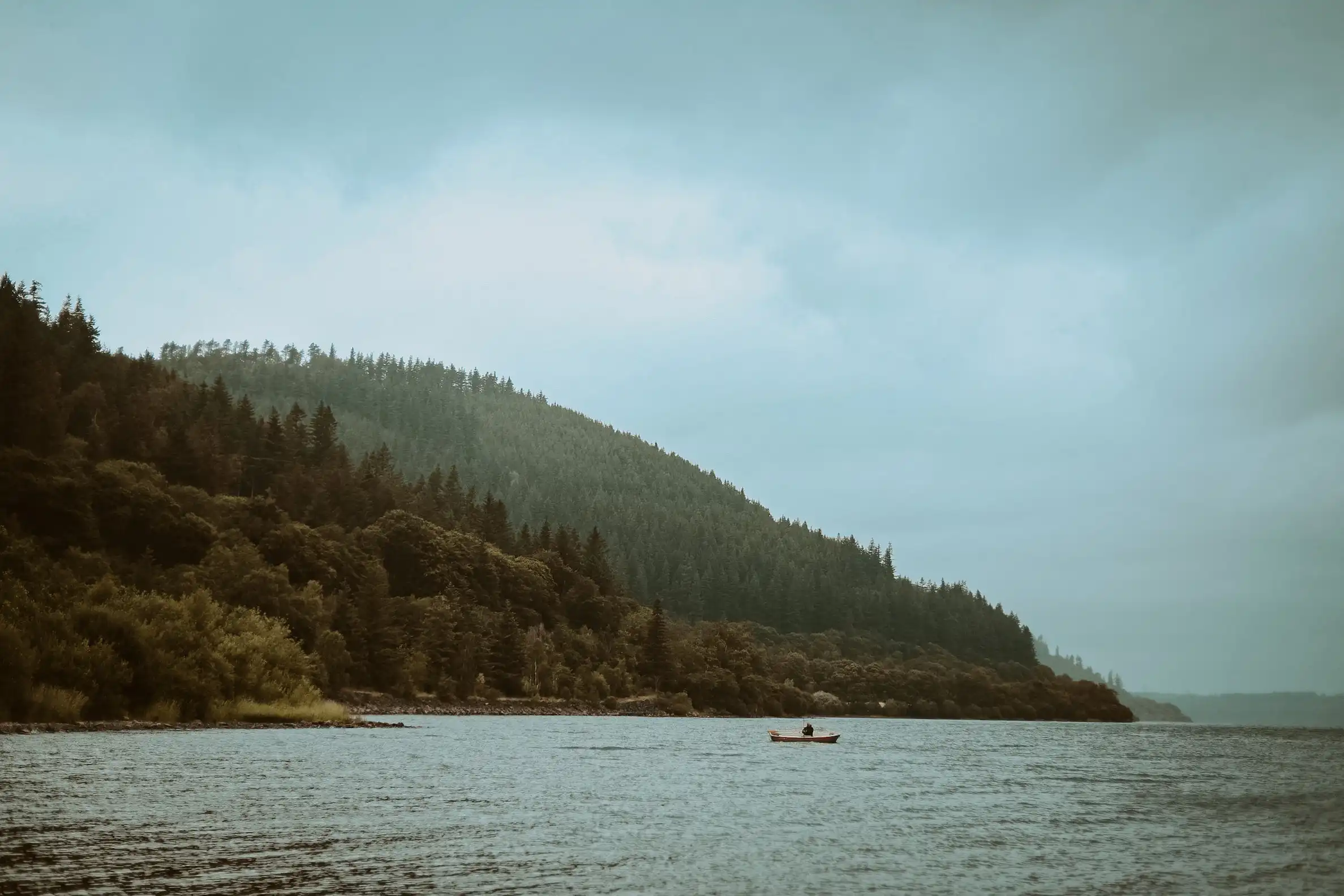 A distant shot of a boat in the middle of a lake. In the background, forests can be seen.