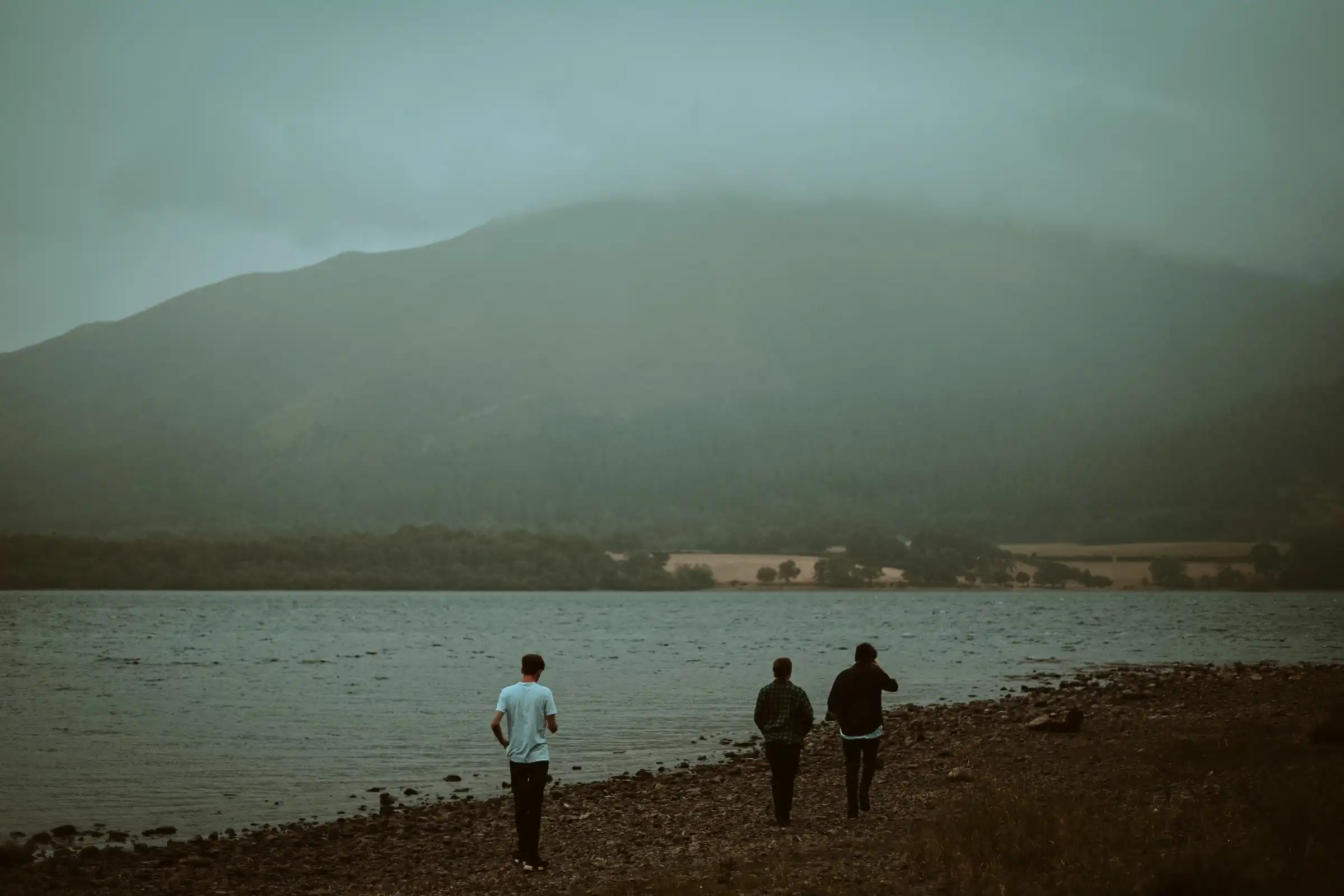 A distant shot of three males walking away from the camera towards a large lake.