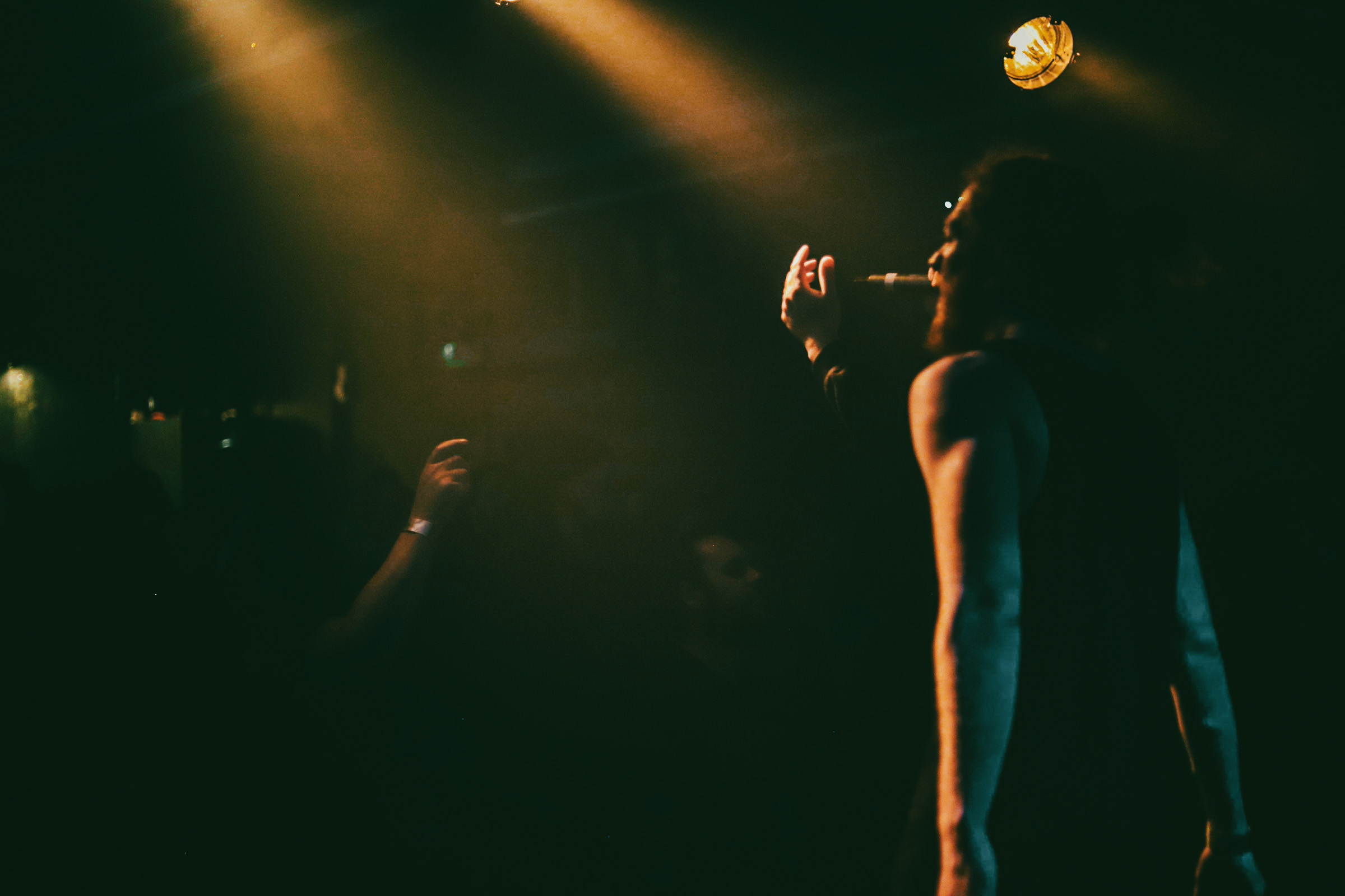 Performers on stage in front of a crowded room, making you feel warm due to lighting, dark room also so not too much more to see.